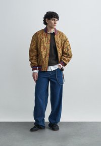 Bomber trapuntato dorato con motivi intricati, polsini a coste in blu e rosso, abbinato a jeans blu larghi e calzature nere.