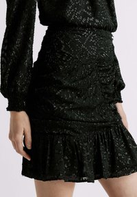 Vestido negro confeccionado en una tela texturizada con un patrón sutil, mangas largas y un dobladillo con volantes. Silueta ajustada.