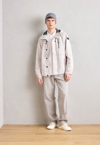 Light gray jacket with black accents, button closures, and adjustable hood, paired with light gray pants and white sneakers. Smooth texture.