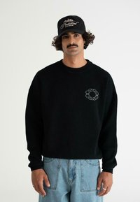 Uomo con baffi e capelli ricci indossa un felpa nera con logo a catena, jeans blu chiaro e cappellino nero con la scritta "Proibito 10119 Berlino".