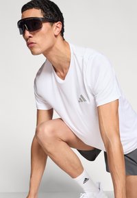 Gafas de sol deportivas Adidas, montura negra, lentes grandes y oscuras. Camiseta blanca que absorbe la humedad, de manga corta, con el logo de Adidas en plata.