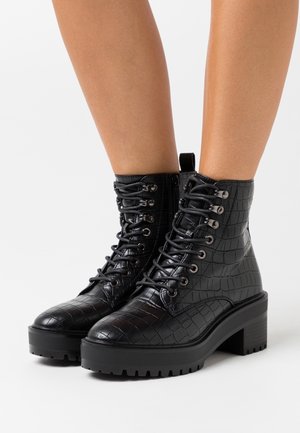 Bottines à lacets en cuir noir effet crocodile avec talons épais, portées sur des jambes nues sur fond blanc.