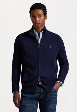Navy zip-up sweater made of knit fabric, featuring a high collar, ribbed cuffs, and a small embroidered logo on the chest. Paired with denim.