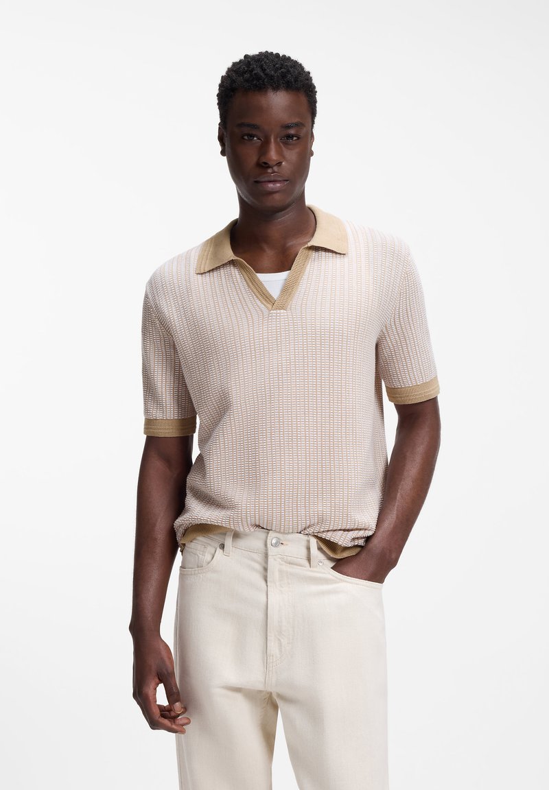 Jeune homme à la peau mate portant un polo à manches courtes en maille beige et un pantalon de couleur claire, debout avec une main dans la poche.