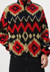 Fleece pullover med halv lynlås, der har et multikolor geometrisk mønster i rød, blå, sort og beige; blød tekstur og afslappet pasform.
