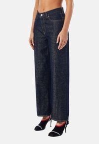 Tmavě modré džíny s širokými nohavicemi a jemnými švy, s klasickým zapínáním na knoflíky a předními kapsami, kombinované s černými podpatky.