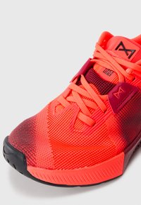 Red athletic shoe with a mesh upper, textured synthetic overlays, and a black rubber sole. Features bright red laces and a contrasting accent.