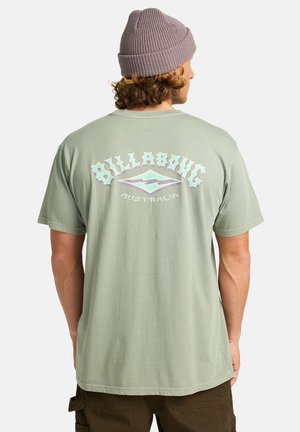 Světle zelené bavlněné tričko s nápisem "Billabong Australia" v pastelových barvách na zádech. Je nošeno s šedou žebrovanou čepicí.
