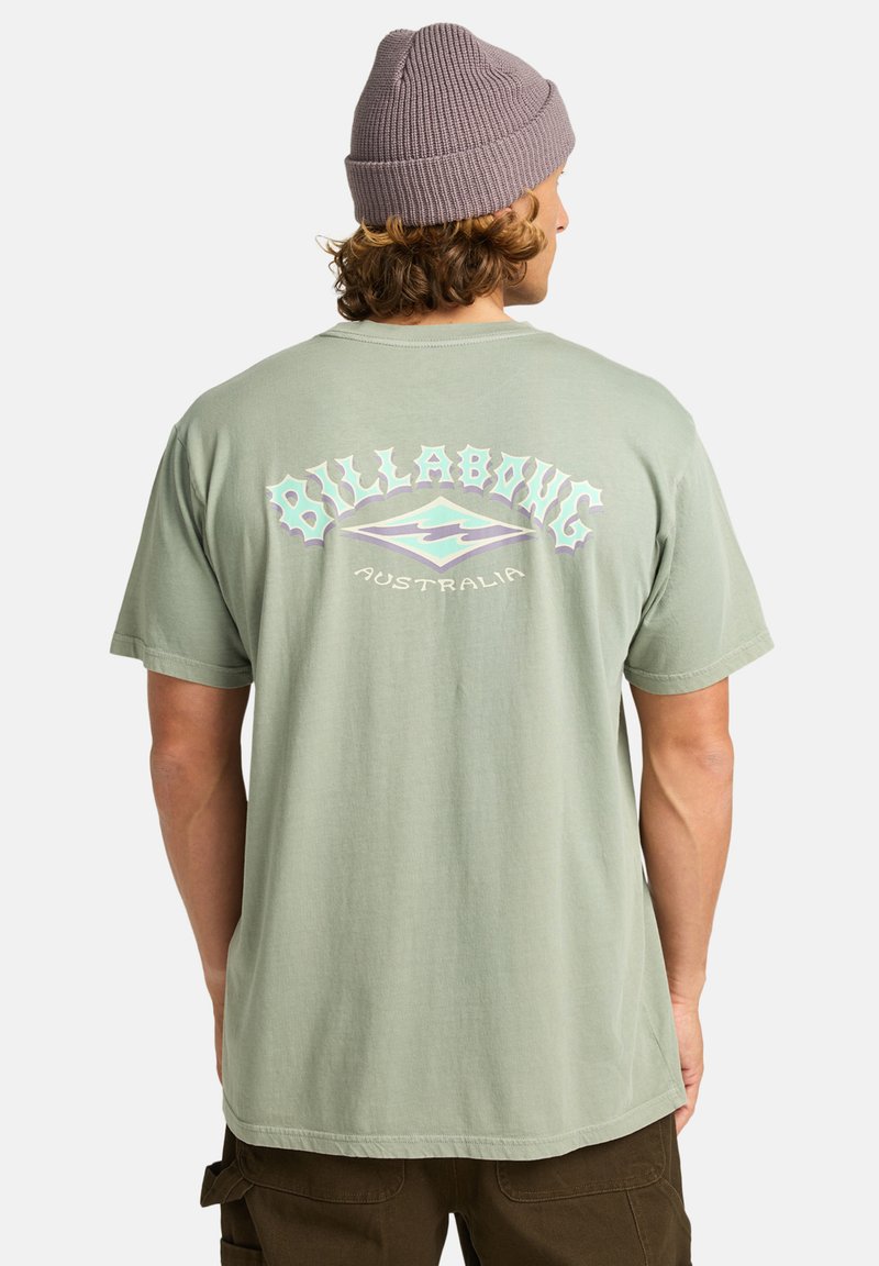 Světle zelené bavlněné tričko s nápisem "Billabong Australia" v pastelových barvách na zádech. Je nošeno s šedou žebrovanou čepicí.
