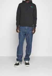 Svart hoodie med en framficka och en logopatch, tillsammans med blå denimjeans med synlig söm och en avslappnad passform. Svarta sneakers med detaljer.