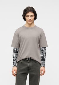 Νεαρό άτομο με κυματιστά σκούρα μαλλιά που φοράει ένα γκρι υπερμέγεθες T-shirt πάνω από μαύρα και λευκά μακριά μανίκια με ζεμπρέ μοτίβο και ξεθωριασμένα μαύρα τζιν.