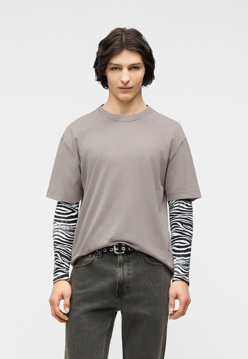 Νεαρό άτομο με κυματιστά σκούρα μαλλιά που φοράει ένα γκρι υπερμέγεθες T-shirt πάνω από μαύρα και λευκά μακριά μανίκια με ζεμπρέ μοτίβο και ξεθωριασμένα μαύρα τζιν.