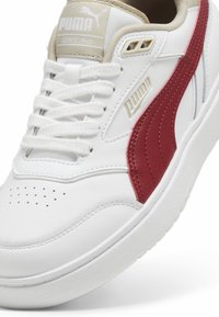 Baskets en cuir blanc avec accents rouges, semelle texturée et détails perforés. Présente un col rembourré et un système de laçage à l'avant.