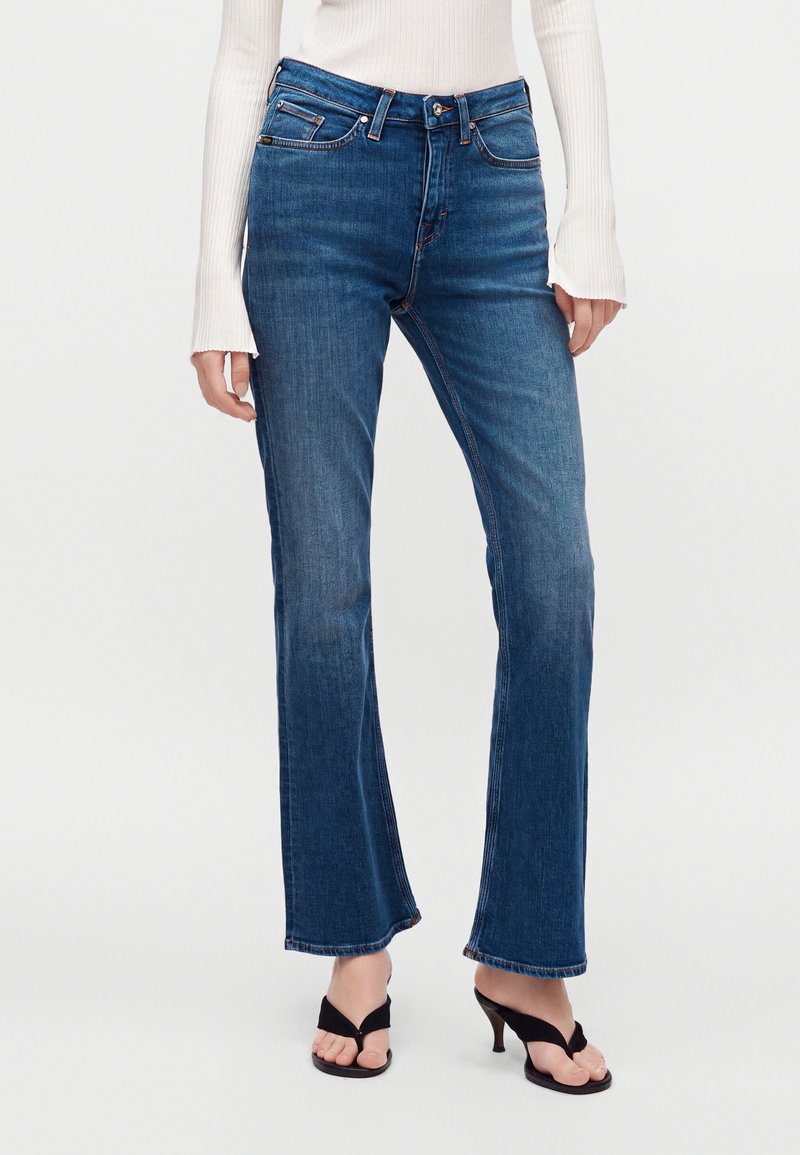 Γυναίκα με μπλε τζιν παντελόνι τύπου bootcut, λευκό τοπ με μακριά μανίκια και μαύρα ανοιχτά σανδάλια με τακούνι, στέκεται μπροστά από απλό φόντο.