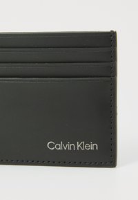 Portofel de carduri din piele neagră, cu multiple sloturi, având un design elegant și logo-ul „Calvin Klein” embosat în colțul din dreapta jos.
