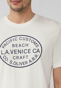 Witte katoenen T-shirt met korte mouwen, voorzien van een marineblauw grafisch ontwerp in een ovale vorm met de tekst "PACIFIC CUSTOMS BEACH L.A. VENICE CA."