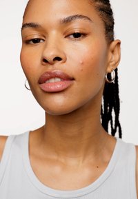 Primo piano di una persona con pelle di colore marrone chiaro che indossa una canottiera grigia. I dettagli notevoli includono piccole imperfezioni sul viso e orecchini a cerchio in argento.