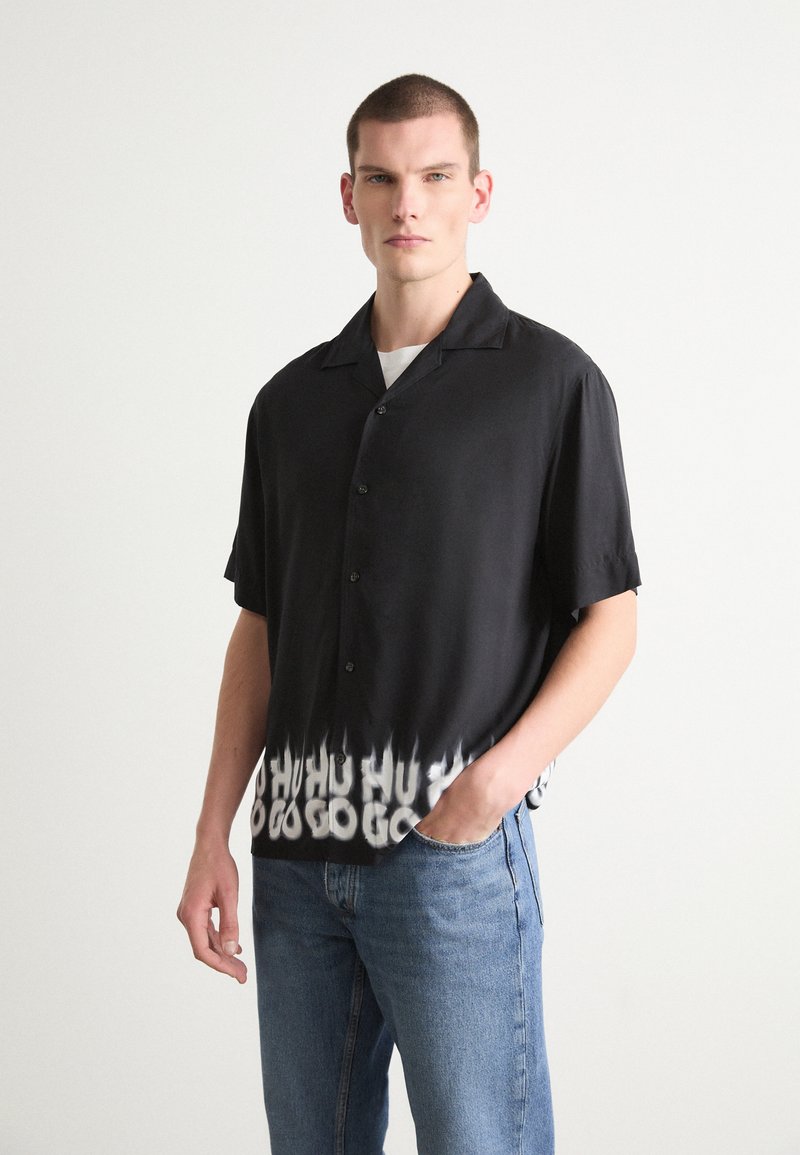 Cămașă neagră cu mâneci scurte, stil cămașă cu nasturi, are un tiv gradient cu un model de text alb. Material neted, croi lejer, purtată cu blugi din denim.