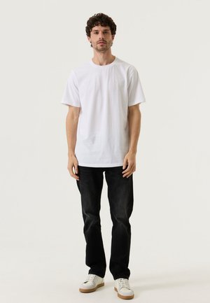 Mann, der ein schlichtes weißes T-Shirt, schwarze Jeans und weiße Sneakers trägt und vor einem neutralen Hintergrund steht.
