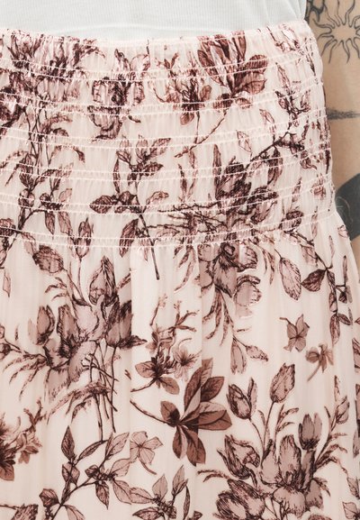 Lyserød nederdel med brunt blomsterprint og elastisk, rynket taljebånd, båret med en hvid top og synlig tatoveret arm i baggrunden.