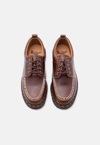 Ζευγάρι καφέ δερμάτινα παπούτσια Dr. Martens με λευκή ραφή, μαύρες σόλες και τέσσερις οπές με σκουρό καφέ κορδόνια, σε θέαση από πάνω.