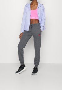 Lys lilla lynlåstrøje over en pink crop-top, grå joggingbukser med rødt logo og sorte sneakers med hvide såler, på en neutral baggrund.