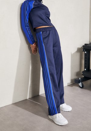 Person in dunkelblauem Trainingsanzug mit blauen Streifen lehnt rückwärts an eine beige Wand und trägt weiße Turnschuhe.