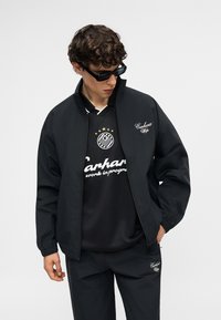 Νεαρός άντρας που φοράει μαύρο μπουφάν Carhartt WIP, ταιριαστό παντελόνι και μαύρο μπλουζάκι με στάμπα, κοιτώντας προς τα κάτω με γυαλιά ηλίου.
