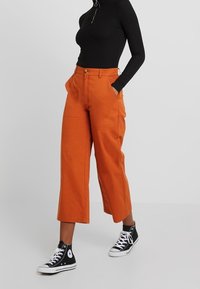 Oranje wijde broek met voorzakken en een knoopsluiting, gecombineerd met een zwarte langemouwen top en zwarte hoge sneakers.