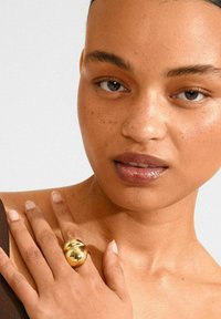 Close-up of a woman with freckles, glossy lips, and a large shiny gold ring on her finger resting on her shoulder.
