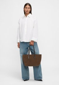 Donna che indossa una camicia bianca button-up larga e jeans a gamba larga, mentre tiene una grande borsa a tracolla marrone con manici neri.