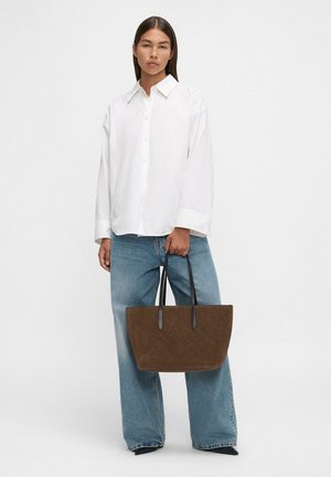 Vrouw in een losse witte blouse met knopen en een wijdere spijkerbroek, die een grote bruine tote bag met zwarte handvatten vasthoudt.