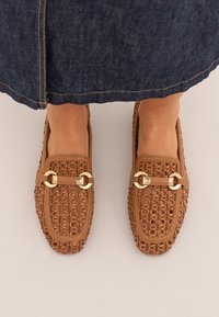 Mocasines marrones trenzados con suela plana, acentos en dorado y un detalle de correa rectangular. Superficie texturizada con tejidos abiertos.