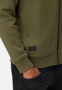 Olijfgroene ritssluitingjasje met een hexagonaal textuurpatroon, ribgebreide boorden en een zwart logo-label aan de onderzijde.