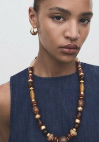 Houten kralenketting met gouden accenten, met verschillende tinten bruin en geel. Model draagt gouden ringen en een denim top.