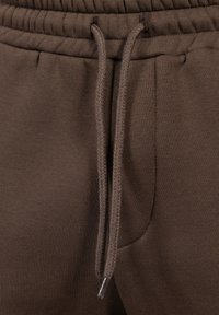 Pantalones de chándal marrones con una cinturilla elástica y cordones, bolsillo lateral externo y una tela suave y texturizada.