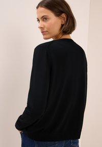 Zwarte gebreide cardigan met lange mouwen en ribbels aan de onderkant. Soepele textuur, eenvoudig ontwerp en een ronde hals, gedragen over blauwe jeans.