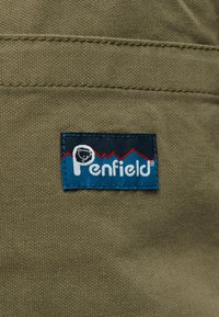 Ετικέτα της μάρκας Penfield ραμμένη σε ελαιοπράσινο ύφασμα με κόκκινο περίγραμμα βουνού και λευκό κείμενο σε μπλε φόντο.