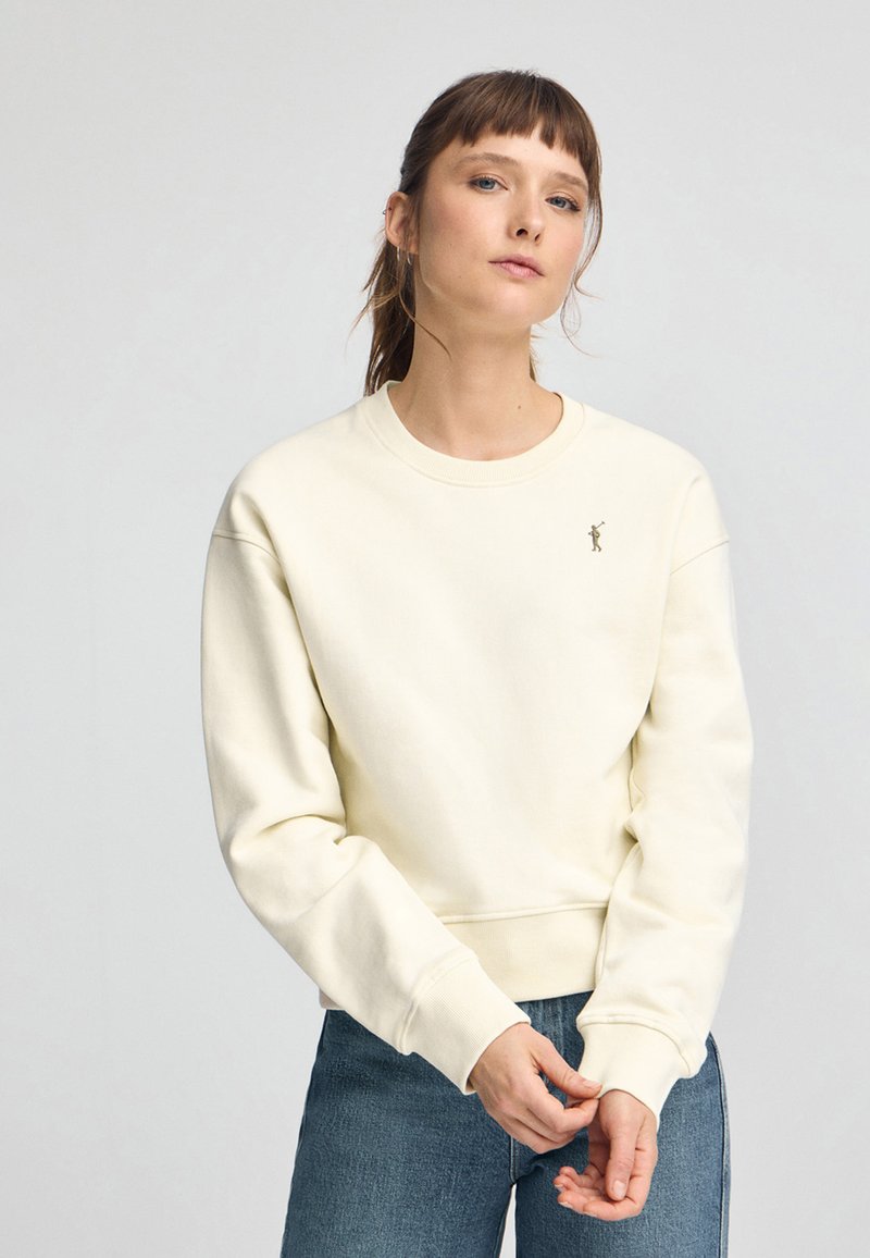 Sudadera beige clara con cuello redondo y puños acanalados. Presenta un pequeño diseño bordado en el pecho. Combinada con jeans azules.