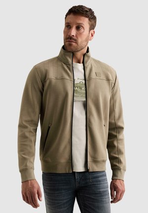 Homme debout portant une veste beige zippée sur un t-shirt blanc à motif graphique et un jean bleu, regardant légèrement sur sa gauche sur un fond uni clair.