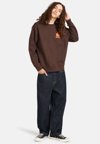 Felpa marrone con un logo arancione sul petto, abbinata a jeans denim scuri larghi e sneakers nere con suole bianche.