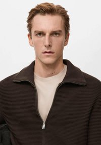 Brązowa kurtka zapinana na zamek z fakturowanym wzorem dzianiny, z kołnierzem i gładką jasną koszulką pod spodem. Tło jest jednolicie białe.