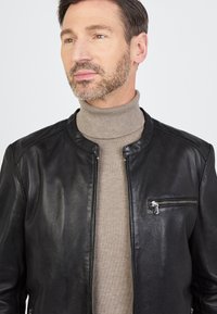 Man met kort bruin haar en een baard, gekleed in een beige coltrui onder een ongeopende zwarte leren jas met een ritszak op de borst.