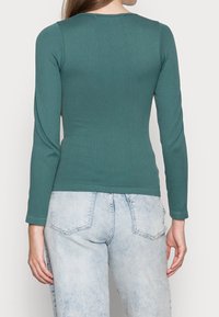 Langärmliges Oberteil aus türkisfarbenem Rippstoff mit Rundhalsausschnitt, taillierter Form und dezenten Nahtdetails. Kombiniert mit hellblauen, ausgebleichten Jeans.