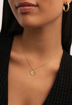 Collar con colgante en forma de corazón dorado sobre una delicada cadena, con una superficie texturizada, llevado sobre la piel con un fondo de tela oscura.