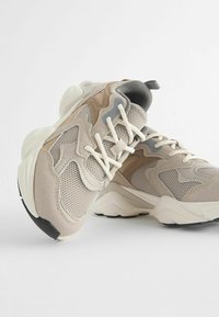 Beige sneakers til sport lavet af mesh og suede materialer, med strukturerede mønstre, cremede snørebånd og en sort gummisål. Elegant silhuet og design.