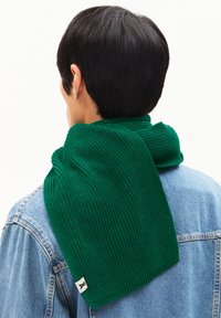 Écharpe en tricot verte présentée enroulée autour du cou. Tissu texturé avec un motif côtelé, comportant une petite étiquette de logo sur le bord.