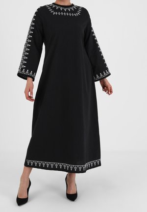 Personne portant une longue robe noire ornée d'une broderie géométrique blanche au niveau du col, des manches et de l'ourlet, associée à des talons noirs pointus.