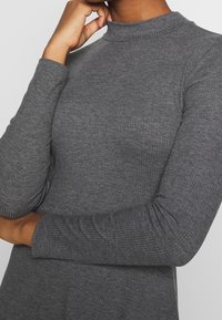 Pull-over gris côtelé à manches longues et col montant. Le tissu présente une texture lisse et une silhouette ajustée, mettant en valeur un design épuré.