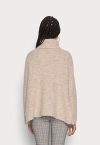 Poncho oversize beige in misto lana con colletto alto, caratterizzato da un motivo a maglia testurizzata e un orlo curvo, indossato sopra pantaloni a fantasia.